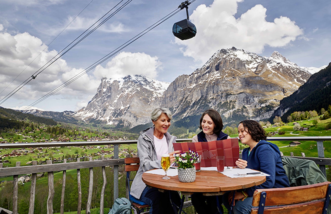 Three women sitting in a restaurant in front of a mountain panorama and a cable car.